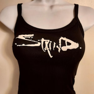 May include: Black ribbed tank top with the word "STAIN" in white, stylized lettering across the front. The tank top has thin straps and a scoop neckline. The garment is displayed on a mannequin.
