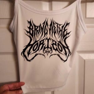 May include: White ribbed tank top with black graphic text that reads "BRING ME THE HORIZON". The top has thin straps and a scoop neckline. The text is in a stylized, gothic font.