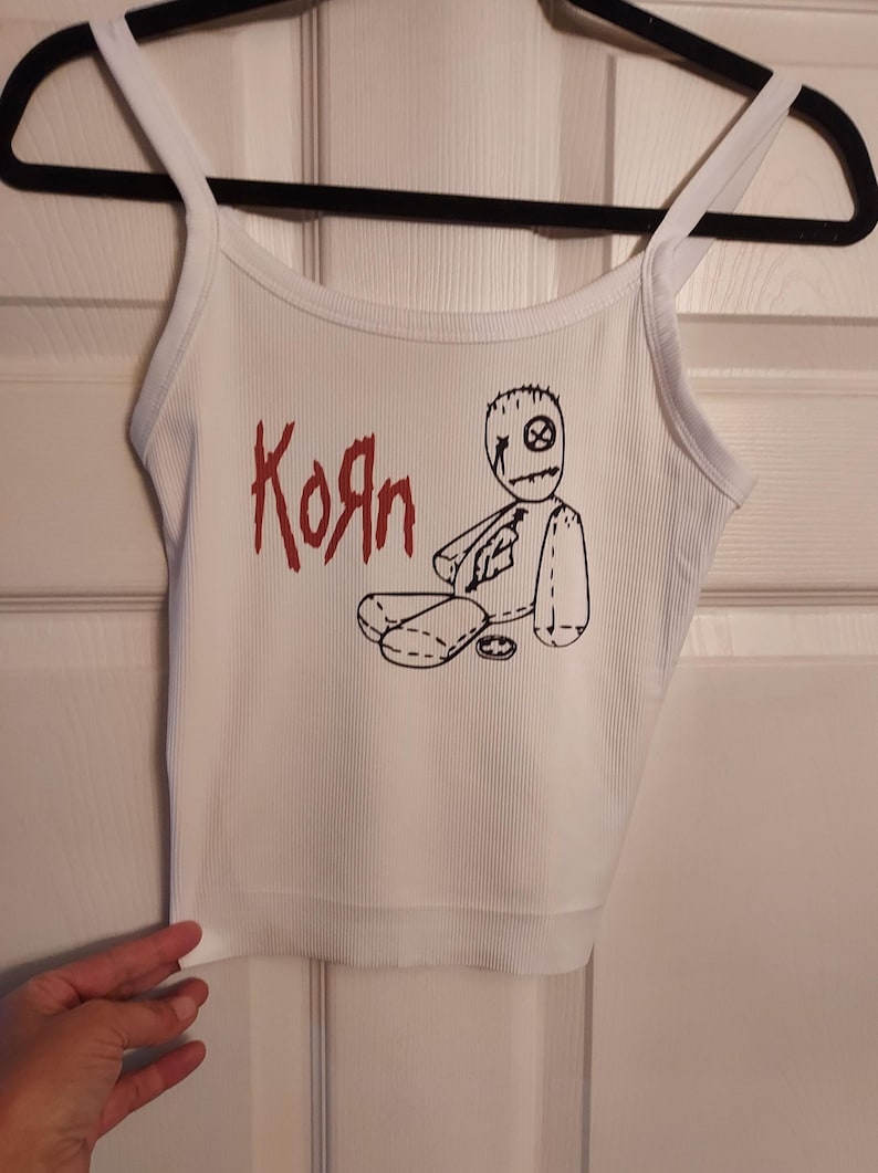 Puede incluir: Camiseta blanca de tirantes con un logotipo rojo de "Korn" y una ilustraci&oacute;n en blanco y negro de una mu&ntilde;eca vud&uacute;.