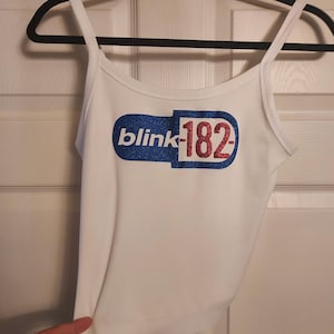 Puede incluir: Camiseta blanca de tirantes con un gr&aacute;fico de una p&iacute;ldora de brillo azul y rojo con el texto "blink-182" en ella.