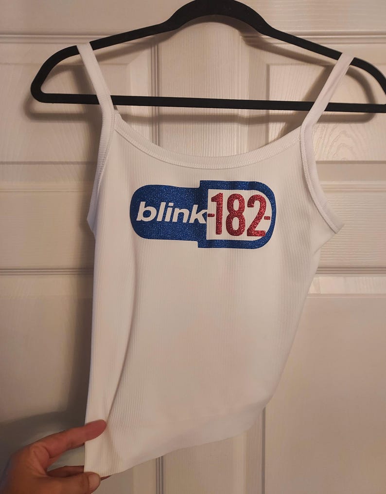 Puede incluir: Camiseta blanca de tirantes con un gr&aacute;fico de una p&iacute;ldora de brillo azul y rojo con el texto "blink-182" en ella.