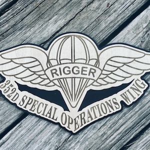 May include: A wooden emblem featuring a parachute and wings design, with the word "RIGGER" on a banner. The emblem also includes the text "352D SPECIAL OPERATIONS WING" in a curved banner shape. The emblem is set against a weathered wood background.