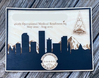 May include: Wooden plaque featuring a skyline silhouette in navy blue, with a light wood background. The plaque includes text: "460th Operational Medical Readiness Sq. May 2022 - Aug 2025" and "SPACE OPERATIONS COMMAND".