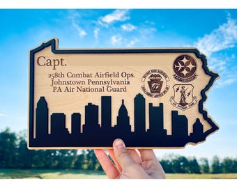May include: Wooden plaque in the shape of Pennsylvania, featuring a black skyline silhouette and text. The plaque includes the words "Capt." and details about the 258th Combat Airfield Ops, Johnstown, PA Air National Guard.