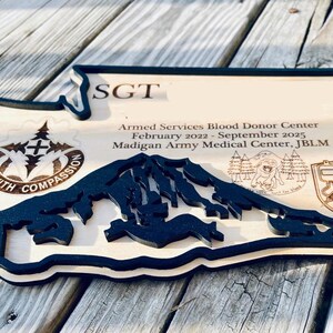 May include: Wooden plaque in the shape of a state, featuring a black mountain range cutout and text. The plaque includes the text "SGT", "Armed Services Blood Donor Center", and "Care with Compassion".