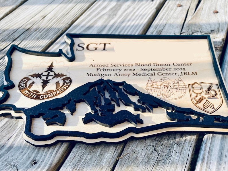 May include: Wooden plaque in the shape of a state, featuring a black mountain range cutout and text. The plaque includes the text "SGT", "Armed Services Blood Donor Center", and "Care with Compassion".