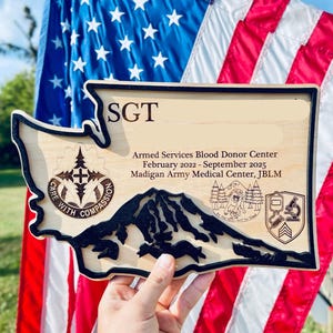 May include: Wooden plaque in the shape of Washington State, featuring a mountain range and text. The plaque reads "SGT" and "Armed Services Blood Donor Center." The background includes an American flag.