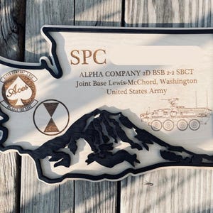 May include: Wooden plaque in the shape of Washington State, featuring engraved text and imagery. The plaque includes the text "SPC", "ALPHA COMPANY 2D BSB 2-2 SBCT", and "Joint Base Lewis-McChord, Washington". It also has a mountain silhouette and military emblems.