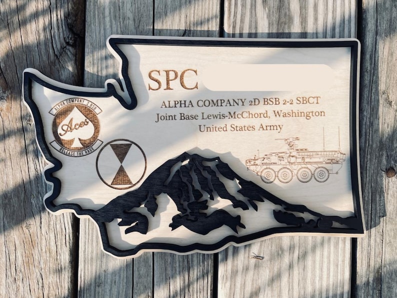 May include: Wooden plaque in the shape of Washington State, featuring engraved text and imagery. The plaque includes the text "SPC", "ALPHA COMPANY 2D BSB 2-2 SBCT", and "Joint Base Lewis-McChord, Washington". It also has a mountain silhouette and military emblems.