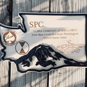 May include: Wooden plaque in the shape of Washington State, with a black border. The plaque features text and graphics, including the words "SPC," "ALPHA COMPANY," and "United States Army." A mountain range and military vehicle are also depicted.