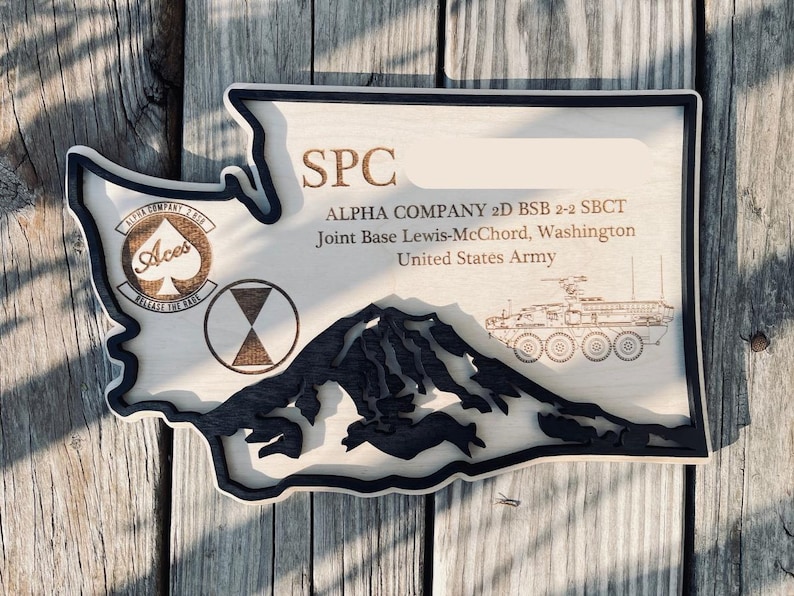 May include: Wooden plaque in the shape of Washington State, with a black border. The plaque features text and graphics, including the words "SPC," "ALPHA COMPANY," and "United States Army." A mountain range and military vehicle are also depicted.