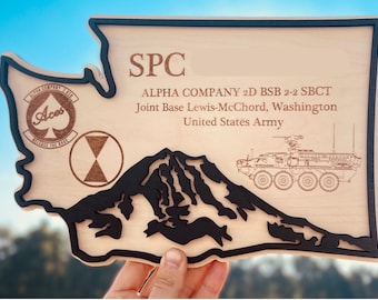 May include: Wooden plaque in the shape of Washington State, with a black border and mountain silhouette. The plaque includes text: "SPC", "ALPHA COMPANY 2D BSB 2-2 SBCT", and "Joint Base Lewis-McChord, Washington, United States Army".