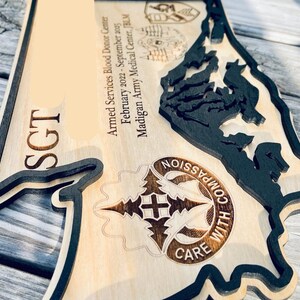 May include: Wooden plaque with a layered design. The plaque features the word "SGT" and text that reads "Armed Services Blood Donor Center February 2022-September 2025 Madigan Army Medical Center, JBLM". The plaque also has a design that says "COMPASSION WITH CARE".