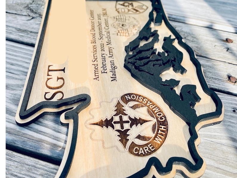 May include: Wooden plaque with a layered design. The plaque features the word "SGT" and text that reads "Armed Services Blood Donor Center February 2022-September 2025 Madigan Army Medical Center, JBLM". The plaque also has a design that says "COMPASSION WITH CARE".