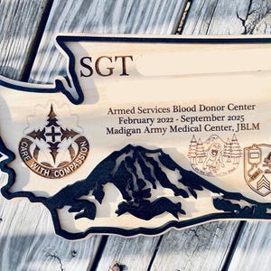 May include: Wooden plaque in the shape of Washington State, featuring engraved text and imagery. The plaque includes the text "SGT", "Armed Services Blood Donor Center", and "February 2022 - September 2025". It also has a mountain range and other military-related emblems.