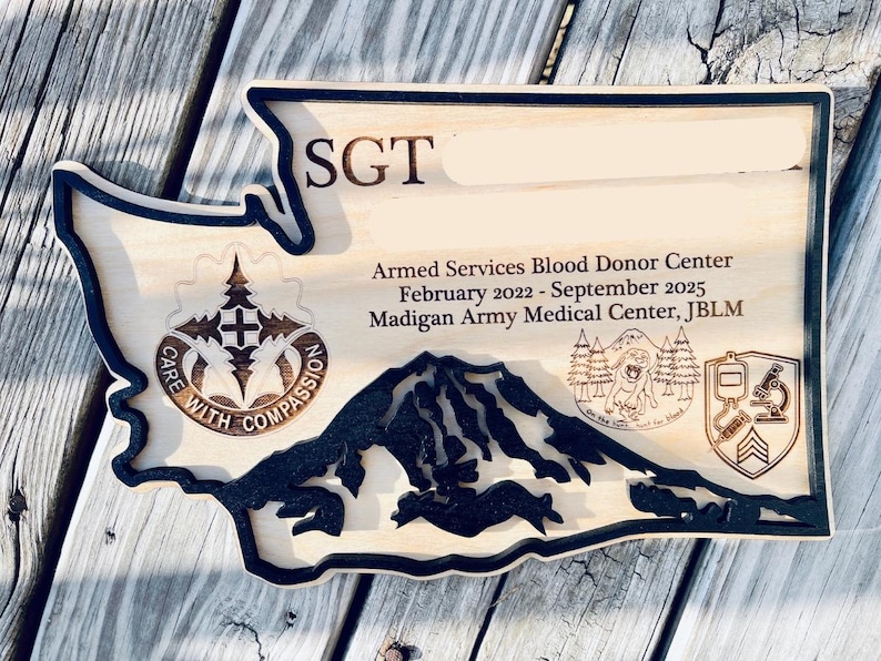 May include: Wooden plaque in the shape of Washington State, featuring engraved text and imagery. The plaque includes the text "SGT", "Armed Services Blood Donor Center", and "February 2022 - September 2025". It also has a mountain range and other military-related emblems.
