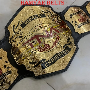 Puede incluir: Un cinturón de campeonato de lucha libre dorado y negro con el texto "WORLD CHAMPION" y "TNA" en rojo. El cinturón tiene detalles intrincados y está hecho de metal y cuero. El texto "HAMYAR BELTS" está impreso en la parte superior.