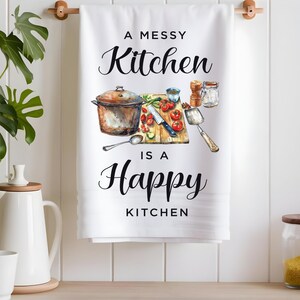 Pode incluir: Toalha de cozinha branca com uma ilustra&ccedil;&atilde;o em preto e branco de uma cozinha desarrumada com o texto "A Messy Kitchen is a Happy Kitchen".