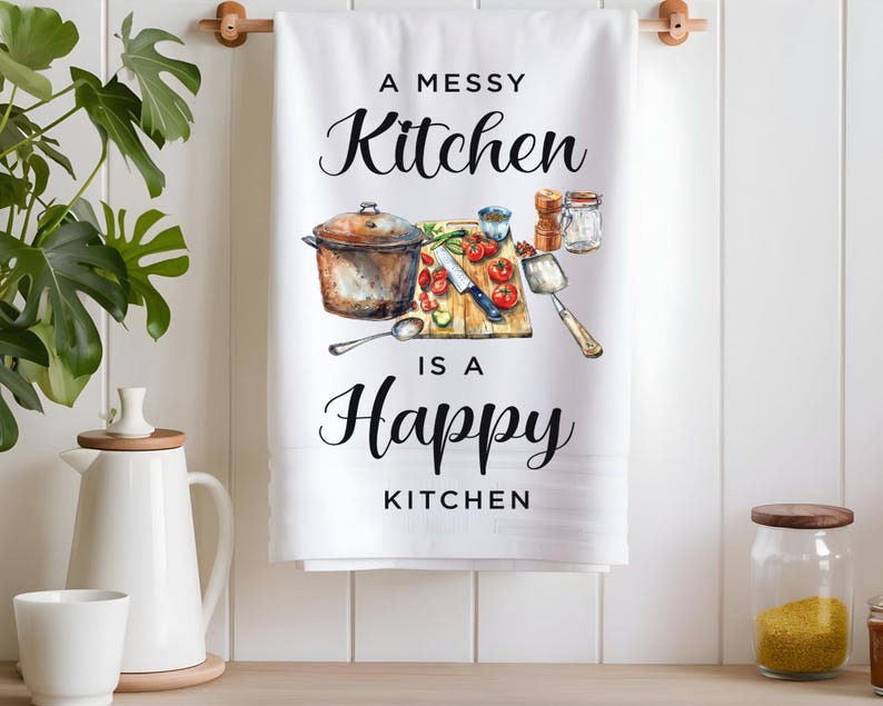 Pode incluir: Toalha de cozinha branca com uma ilustra&ccedil;&atilde;o em preto e branco de uma cozinha desarrumada com o texto "A Messy Kitchen is a Happy Kitchen".