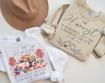 May include: Two white sweatshirts with a fall harvest theme. The text on the sweatshirts reads "The Harvest is plentiful, but the laborers are few." One sweatshirt has a pink truck with pumpkins in the bed. The other sweatshirt has a floral design with pumpkins.