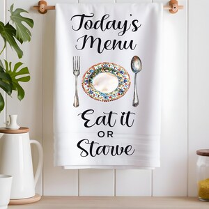 Pode incluir: Toalha de cozinha branca com texto preto que diz "Today's Menu Eat it or Starve". Um prato branco com um design floral colorido &eacute; mostrado com um garfo e uma colher.