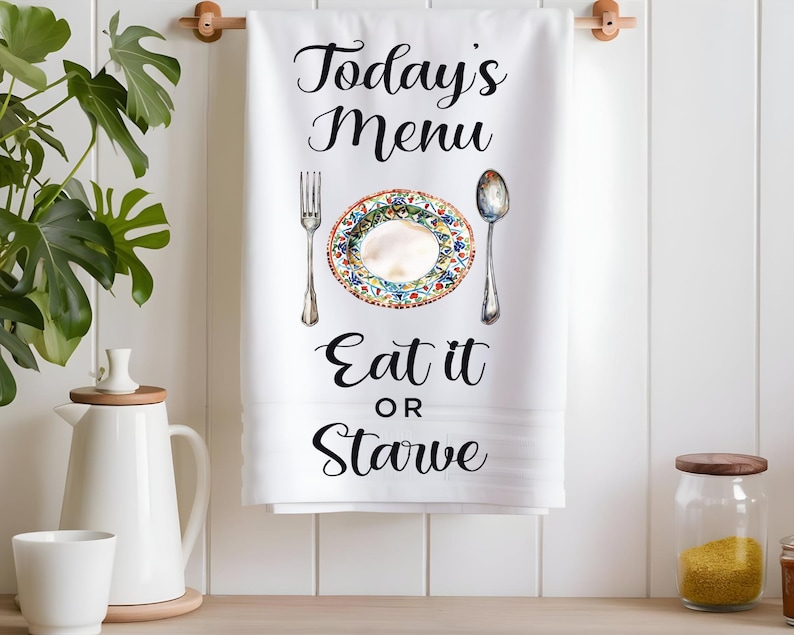 Pode incluir: Toalha de cozinha branca com texto preto que diz "Today's Menu Eat it or Starve". Um prato branco com um design floral colorido &eacute; mostrado com um garfo e uma colher.