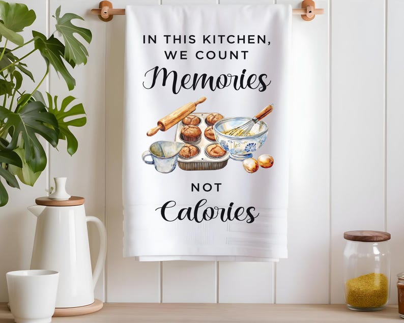 Pode incluir: Toalha de cozinha branca com o texto "Nesta cozinha, contamos mem&oacute;rias, n&atilde;o calorias" e uma ilustra&ccedil;&atilde;o em aquarela de uma assadeira com muffins, uma tigela com um batedor e uma x&iacute;cara medidora.