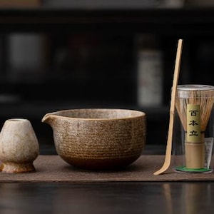Puede incluir: Un juego de té matcha con un cuenco de cerámica, un batidor, una cuchara y un pequeño recipiente de cerámica. El juego es en tonos marrones y beige, con una estética natural y terrosa. El batidor está en un recipiente transparente con texto japonés.