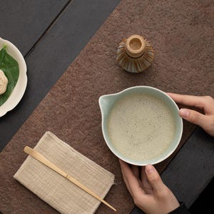 May include: A light green ceramic bowl filled with frothy matcha tea, held by hands on a brown textured surface. A small wooden whisk and a white plate with a green leaf are also visible.