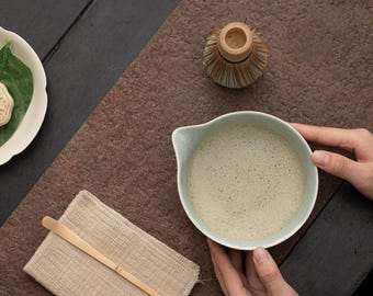 May include: A light green ceramic bowl filled with frothy matcha tea, held by hands on a brown textured surface. A small wooden whisk and a white plate with a green leaf are also visible.