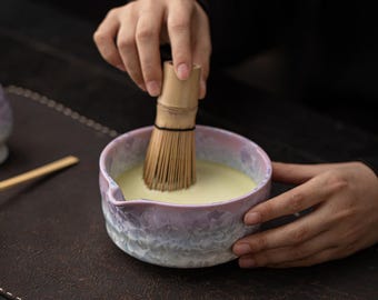 May include: A ceramic matcha bowl with a textured, iridescent glaze in shades of lavender, pink, and white, featuring a small pouring spout. A bamboo matcha whisk is submerged in the pale green liquid inside the bowl, which has a rounded shape.