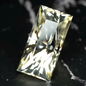 May include: A rectangular, faceted gemstone with a pale yellow, translucent appearance. The stone features a precise, geometric cut with multiple triangular and trapezoidal facets that create sharp light reflections and internal brilliance throughout.