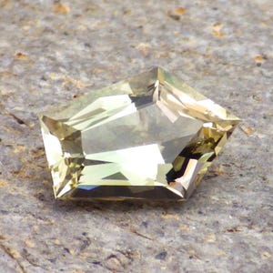 以下が含まれることがあります： 独特な幾何学的な形をした、面取りされた薄い黄色の宝石。石は、尖った頂上と平らな底を持つ五角形にカットされています。半透明で、わずかに輝いています。