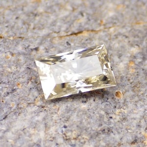 May include: A clear, rectangular, faceted gemstone with a light yellow hue. The gemstone is set against a rough, gray background.