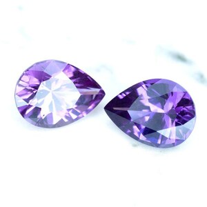 May include: Two pear-shaped amethyst gemstones, one with a lighter purple hue and the other with a darker purple hue.