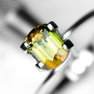 May include: A close-up of a green and yellow gemstone set in a silver ring. The gemstone is cut in an emerald shape.
