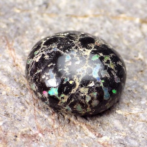 May include: A black opal cabochon with iridescent green, blue, and pink flashes. The opal has a rough, textured surface.