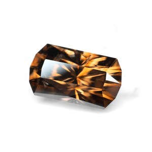 May include: A faceted, rectangular gemstone with a warm, amber color and intricate internal patterns. The gem has a polished surface and a clear, reflective area in the center. The cut of the stone creates a play of light and shadow.