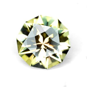 May include: A faceted gemstone with a complex, multi-faceted design. The gem features a blend of pale green and clear facets, creating a play of light and shadow. The gemstone is set against a white background.