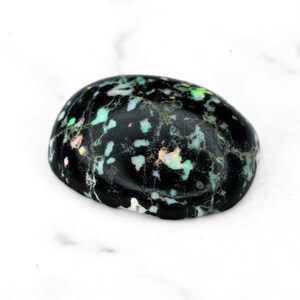 May include: A polished, oval-shaped gemstone with a black base color. The stone is speckled with patches of white, green, and pink, creating a unique, iridescent pattern. The gemstone is smooth and rounded.