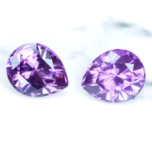 May include: Two pear-shaped purple gemstones with a faceted cut. The gemstones are set against a white marble background.