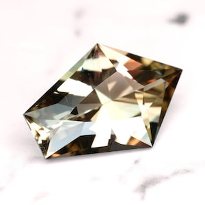 Puede incluir: Una gema facetada de forma rectangular con esquinas anguladas. La gema presenta tonos marrones claros y dorados, con múltiples superficies reflectantes. La gema se encuentra sobre un fondo blanco.