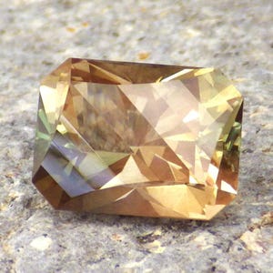Könnte beinhalten: Ein facettierter, goldbrauner Edelstein mit grünen und braunen Farbtönen. Der Edelstein ist in einer rechteckigen Form mit scharfen Kanten und einer glatten, polierten Oberfläche geschliffen.