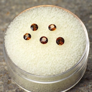 May include: Five round, faceted, dark orange gemstones are displayed in a clear plastic container with a white foam insert. The gemstones have a brilliant cut and are arranged in a scattered pattern. The container sits on a textured, gray surface.