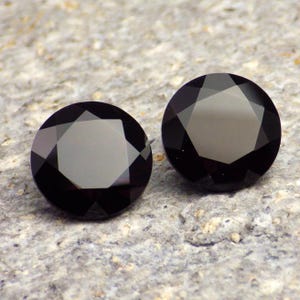 May include: Two black round gemstones with a faceted cut. The gemstones are set against a gray background.