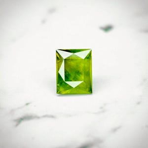 Puede incluir: Una piedra preciosa verde con un corte cuadrado y un diseño facetado.