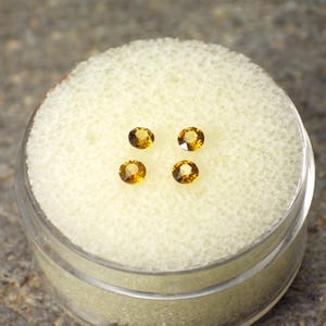 May include: Four oval-cut, golden-yellow gemstones are displayed in a clear plastic container with a white foam insert. The gems are evenly spaced and appear to be faceted, catching the light. The container is partially visible, set against a neutral background.