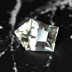 May include: A faceted, clear gemstone with a unique pentagonal shape. The gem has multiple reflective surfaces, creating a sparkling effect. The stone is set against a black and white patterned background, highlighting its clarity and brilliance.