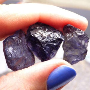 Peut inclure: Trois pierres précieuses naturelles, brutes, de couleur violet foncé. Les pierres sont de forme irrégulière et ont un léger éclat métallique.