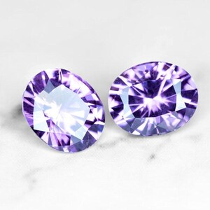 May include: Two oval-shaped amethyst gemstones with a purple and white color gradient. The gemstones are faceted and have a high shine.